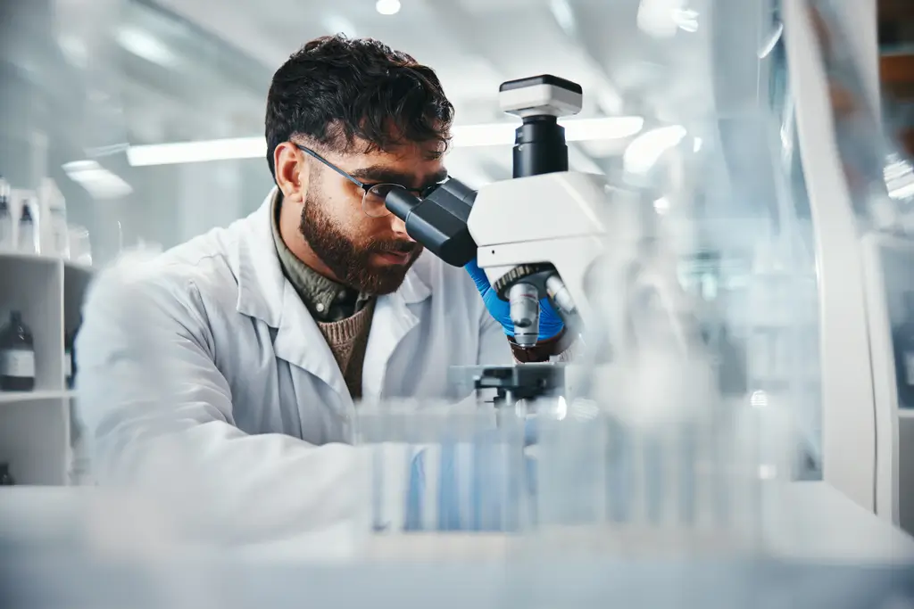 A man in a lab coat examines a sample through a microscope in a laboratory setting.